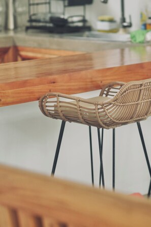 Una silla de mimbre con asiento y respaldo trenzados, sostenida por patas de metal negro, ubicada junto a una mesa de madera.