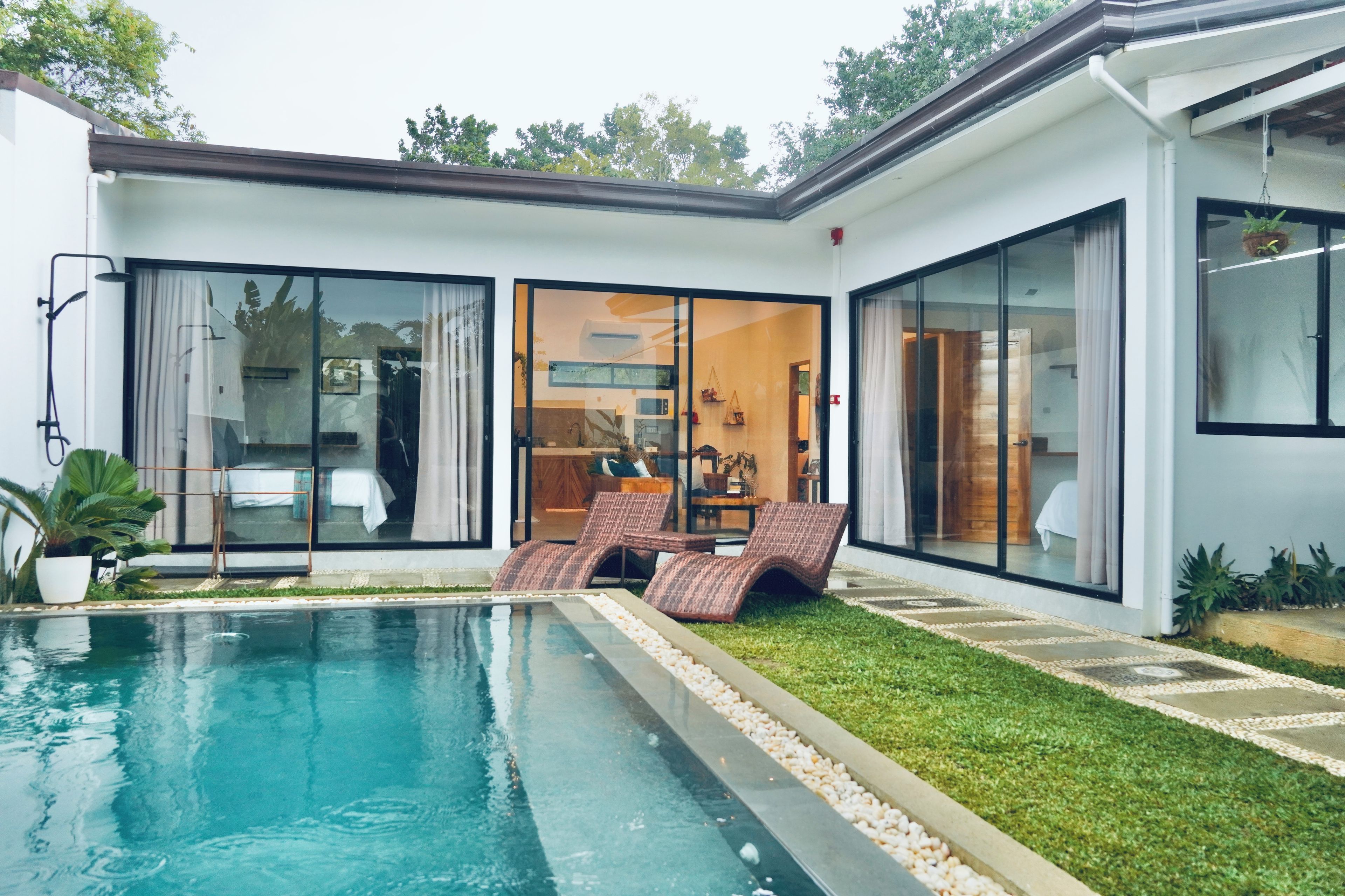 Una casa moderna con piscina, un ventanal que da a una sala de estar, y un césped bien cuidado.