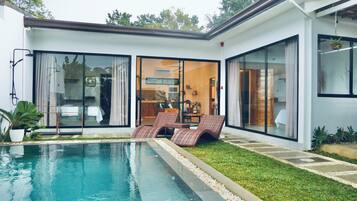 Una casa moderna con piscina, un ventanal que da a una sala de estar, y un césped bien cuidado.