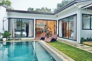 Una casa moderna con piscina, un ventanal que da a una sala de estar, y un césped bien cuidado.