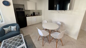 Un espace de vie compact comprenant un coin cuisine, un coin repas avec une table ronde et quatre chaises, un canapé avec des coussins à motifs et un téléviseur à écran plat fixé au mur.