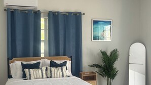 Une chambre à coucher comprenant un lit, des rideaux, un ventilateur de plafond, un climatiseur, une petite table de chevet, une plante en pot, un tableau encadré et une armoire blanche.