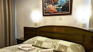 Ein ordentlich bezogenes Bett mit floraler Bettwäsche und zwei gefalteten Handtüchern, ein Frauenbild an der Wand und ein Nachttisch mit einer Lampe.
