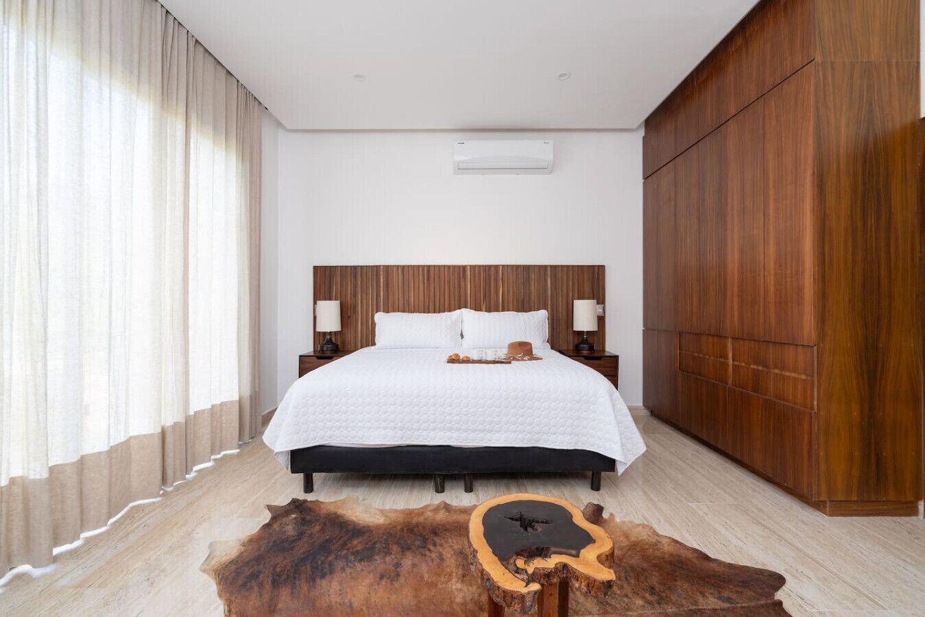 Une chambre d’hôtel moderne avec une tête de lit en bois, un grand lit avec une literie blanche, deux tables de chevet avec des lampes et une armoire en bois.