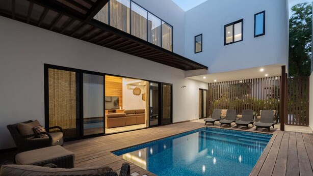 A modern outdoor pool area with a wooden deck, lounge chairs, and a glass-enclosed living space. - Villa Amalia, 5 bdr w/Private Pool in Aldea  Zama (Tulum)