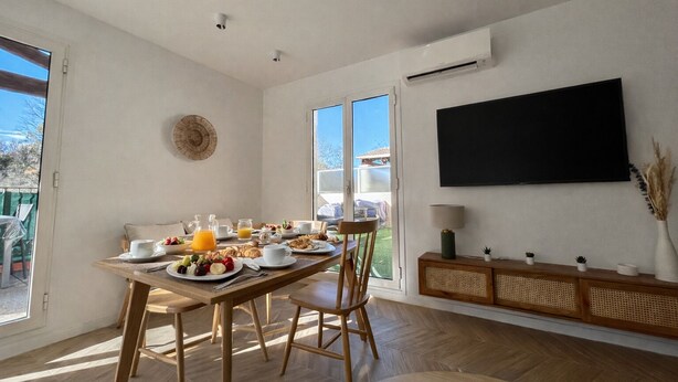 A modern dining area with a wooden table set for breakfast, a large window, and a mounted TV. - Charming apartment with terrace, village on foot (Valbonne)