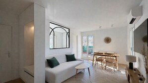 A modern living room with a white sofa, a small round table, and a dining area with wooden chairs. - Charming apartment with terrace, village on foot (Valbonne)