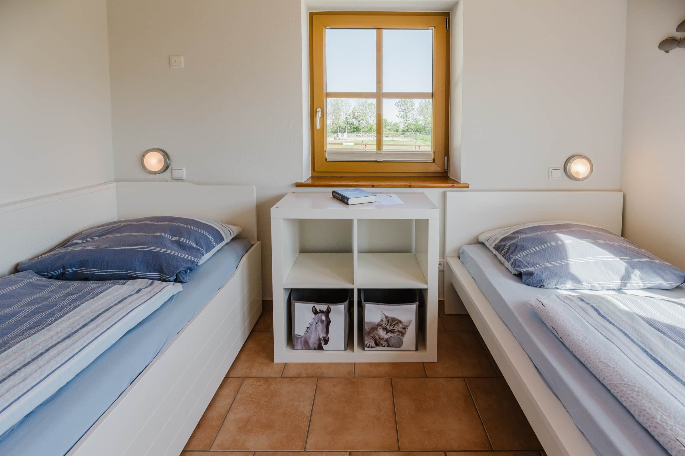 Una habitación pequeña con dos camas, una ventana, un escritorio con espacio de almacenamiento y luces fijadas a la pared.