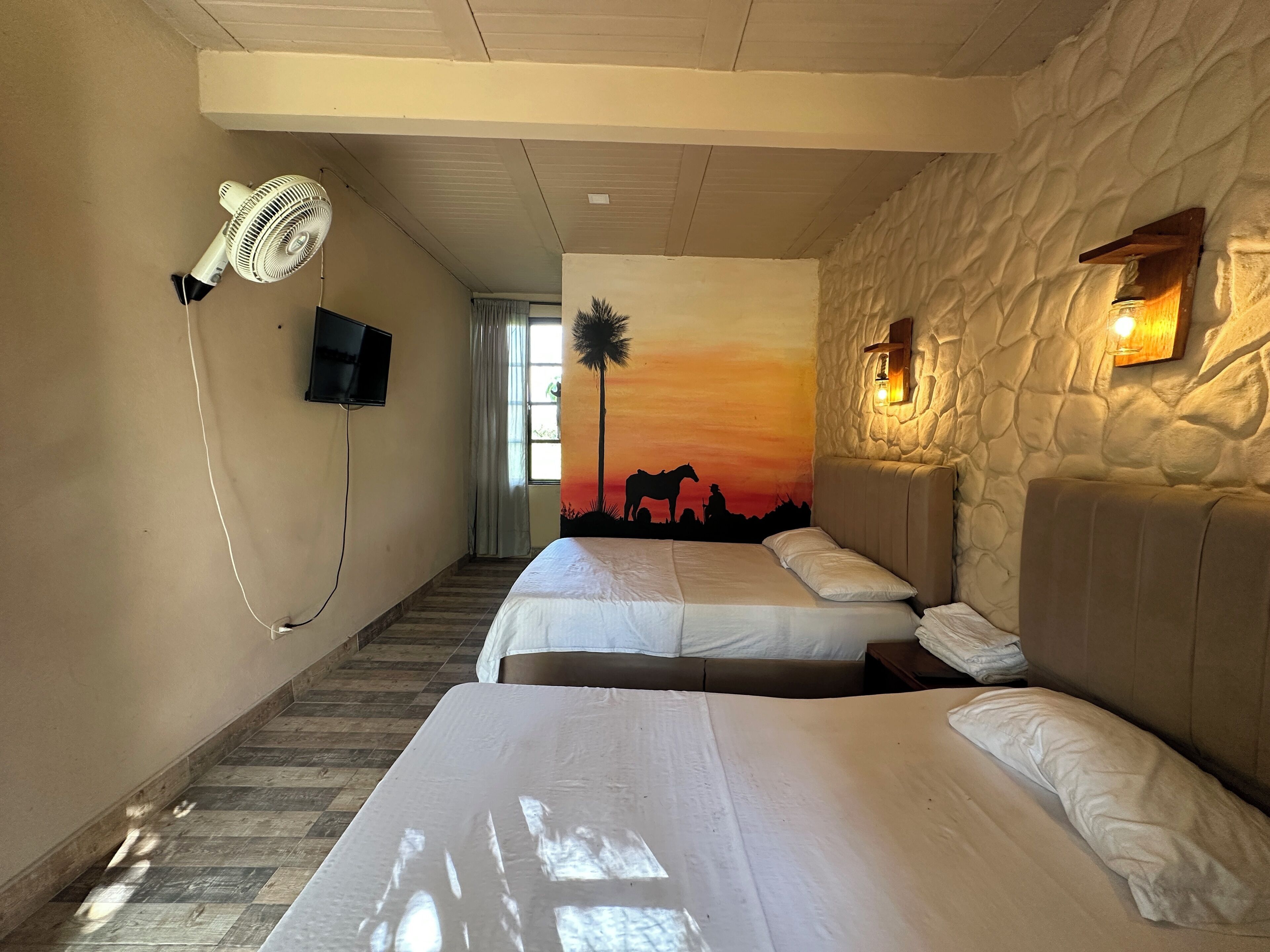 A hotel room with two beds, a ceiling fan, a TV, a sunset mural, and a stone wall.