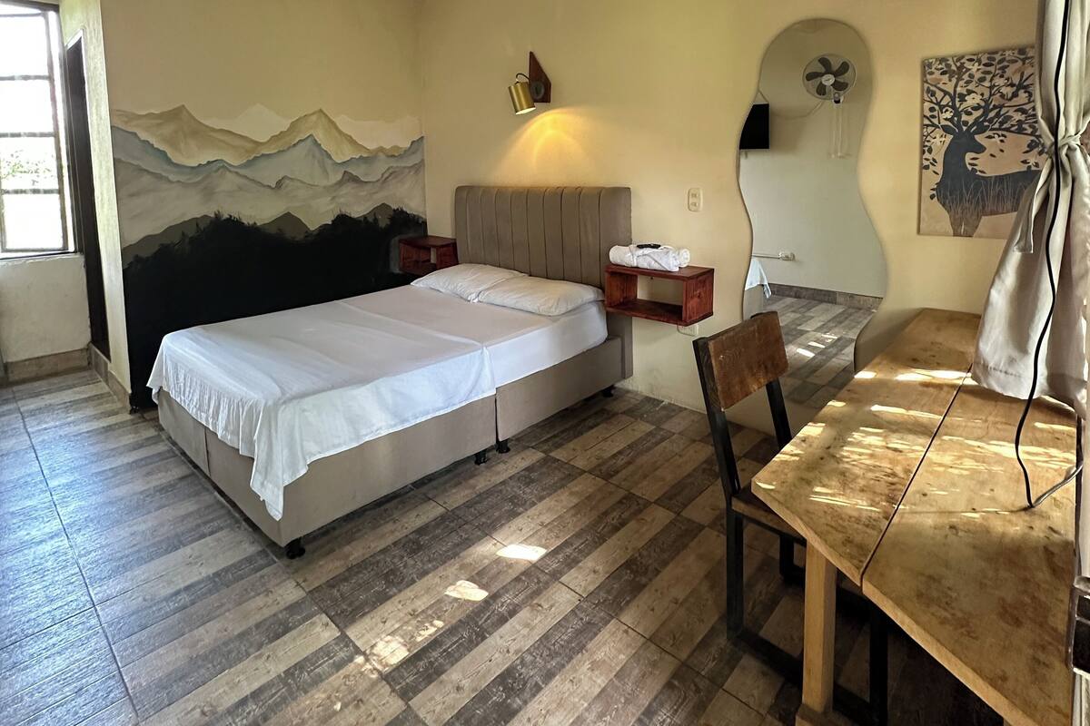 A bedroom with a bed, a desk, a chair, a wall-mounted lamp, and a painting.