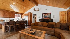 A spacious living area with a wooden ceiling, a large sofa, a coffee table, and a kitchen area with wooden cabinets and a dining table. - Lakeside Cottage, Charming 3-BDRM Retreat W Hottub (Big Bear Lake)
