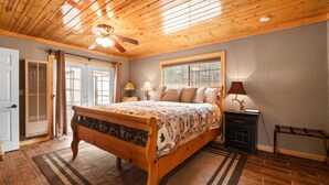 A bedroom with a wooden ceiling, a bed with a patterned bedspread, two bedside lamps, and a window with a view of greenery. - Lakeside Cottage, Charming 3-BDRM Retreat W Hottub (Big Bear Lake)