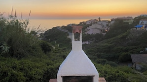 Una estructura de piedra blanca con chimenea, rodeada de vegetación y con vistas a un pueblo costero al atardecer.