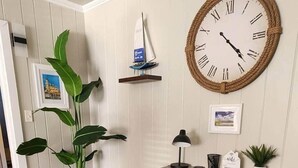 A room with a wooden desk, a potted plant, a wall-mounted clock, a small shelf with a model sailboat, and framed pictures.