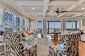 A spacious living room with a view of the ocean, featuring wicker furniture, a ceiling fan, and large windows.