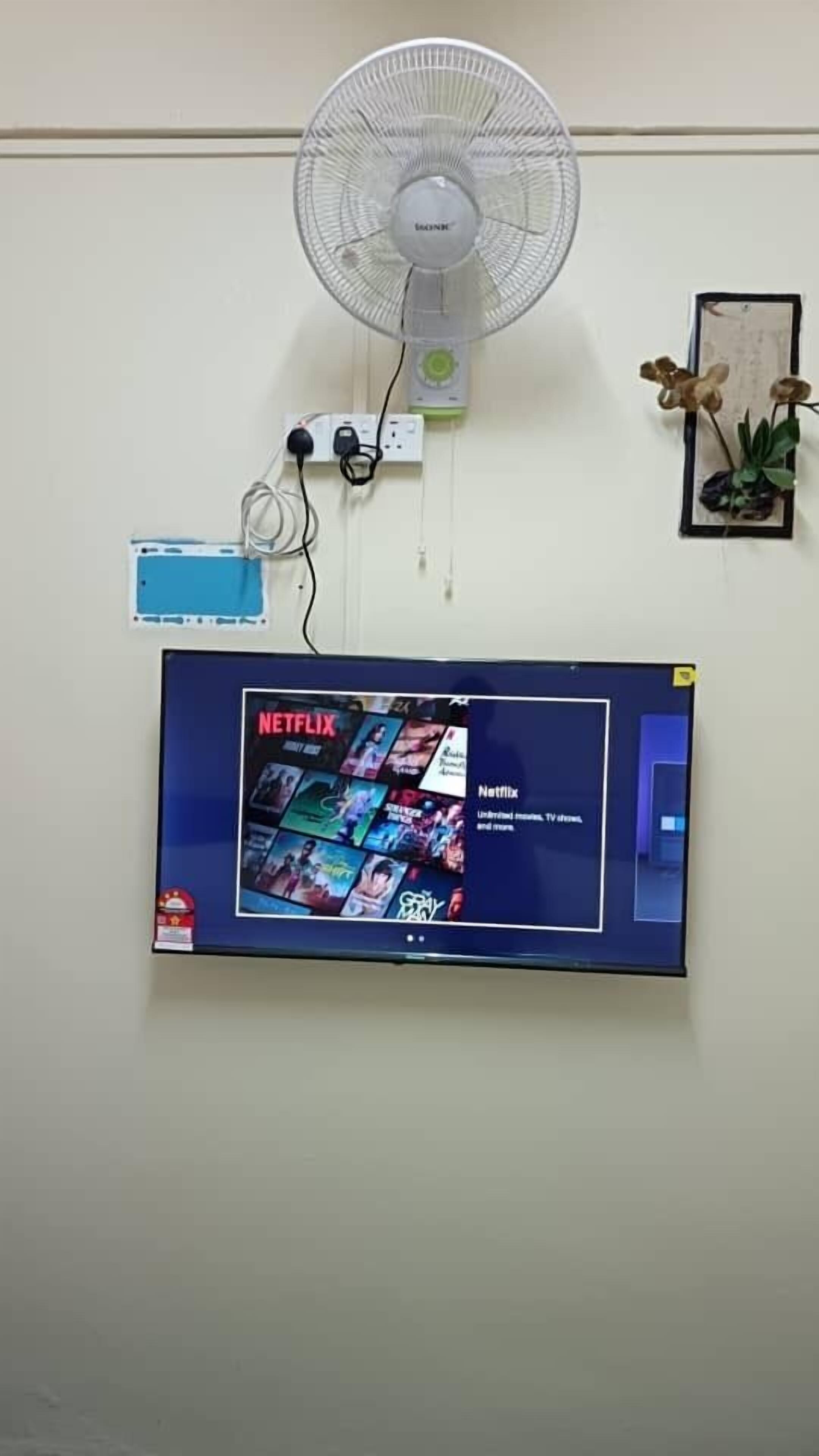 Una TV a parete con contenuti Netflix, una ventola a parete e una ciabatta con cavi.