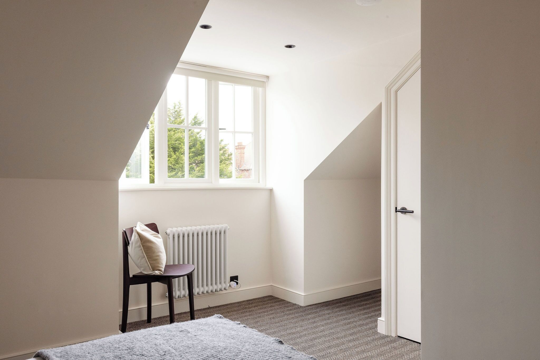 A room with a sloped ceiling, a radiator, a chair, a door, and a window with a view of greenery.
