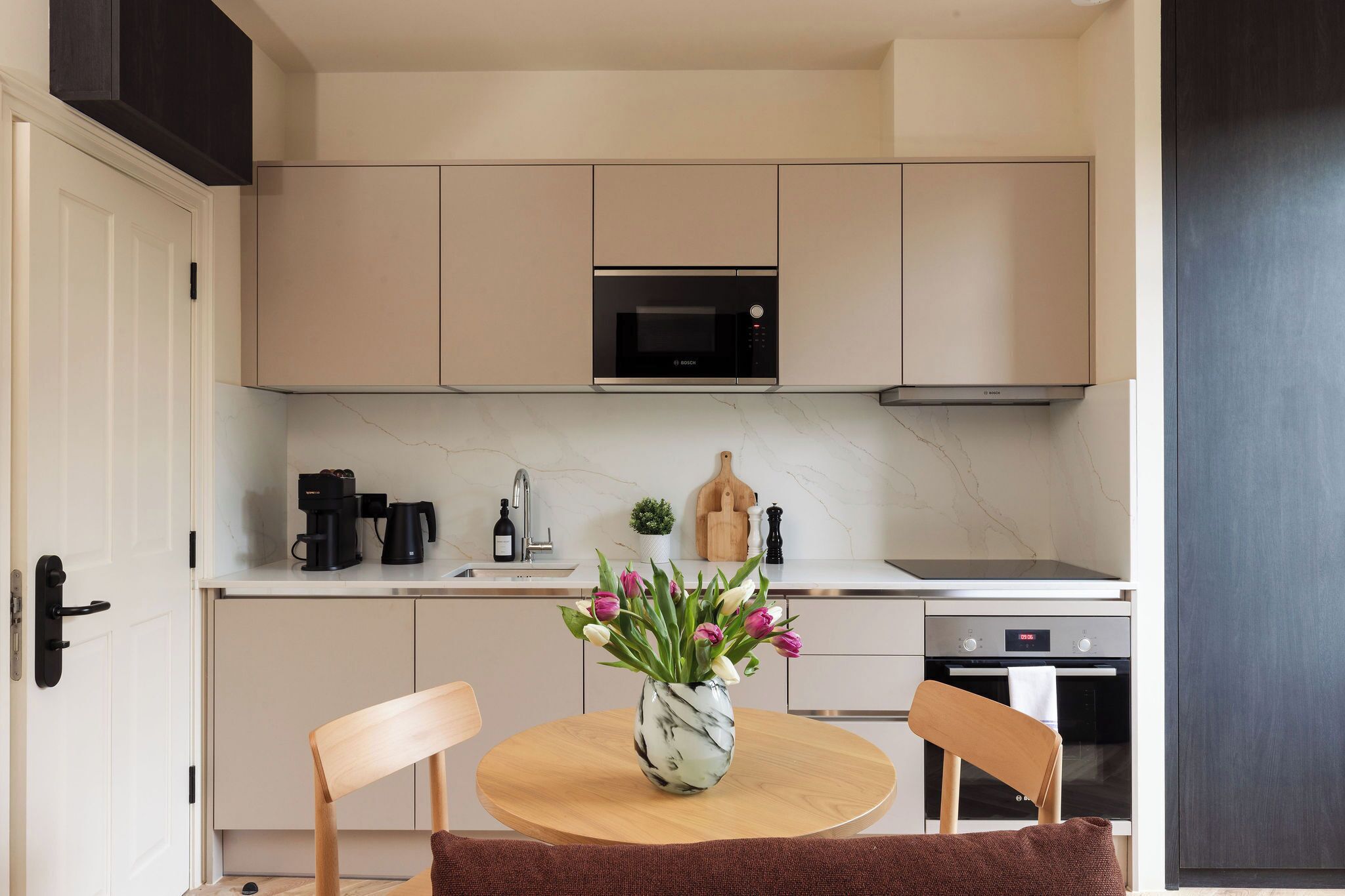 Een moderne keuken met een ingebouwde magnetron, oven en koffiezetapparaat. Er staat een ronde tafel met stoelen en een vaas met bloemen.