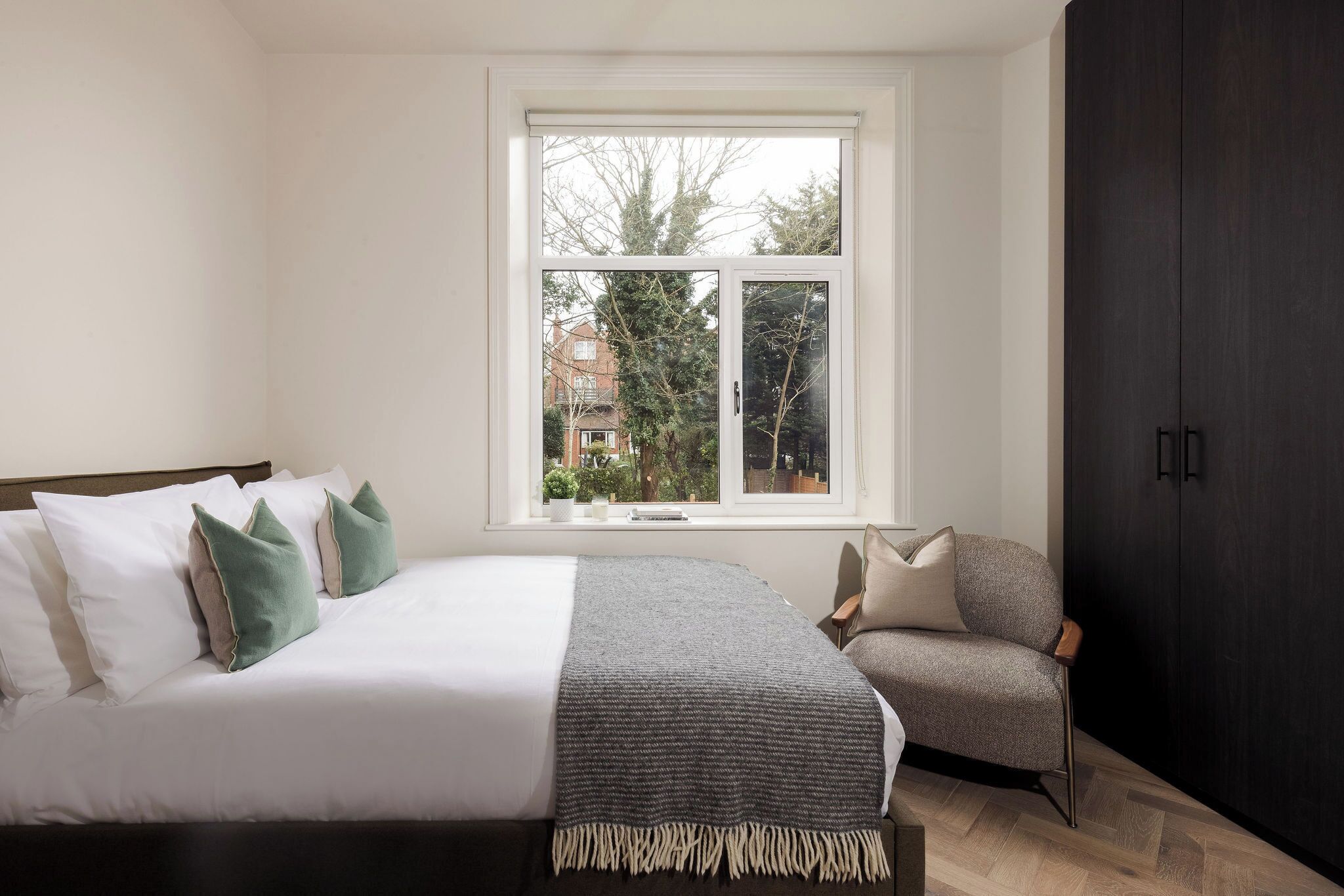 Ein Schlafzimmer mit Bett, Sessel, Kleiderschrank und Fenster mit Blick auf Bäume und Gebäude.
