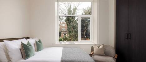 Ein Schlafzimmer mit Bett, Sessel, Kleiderschrank und Fenster mit Blick auf Bäume und Gebäude.