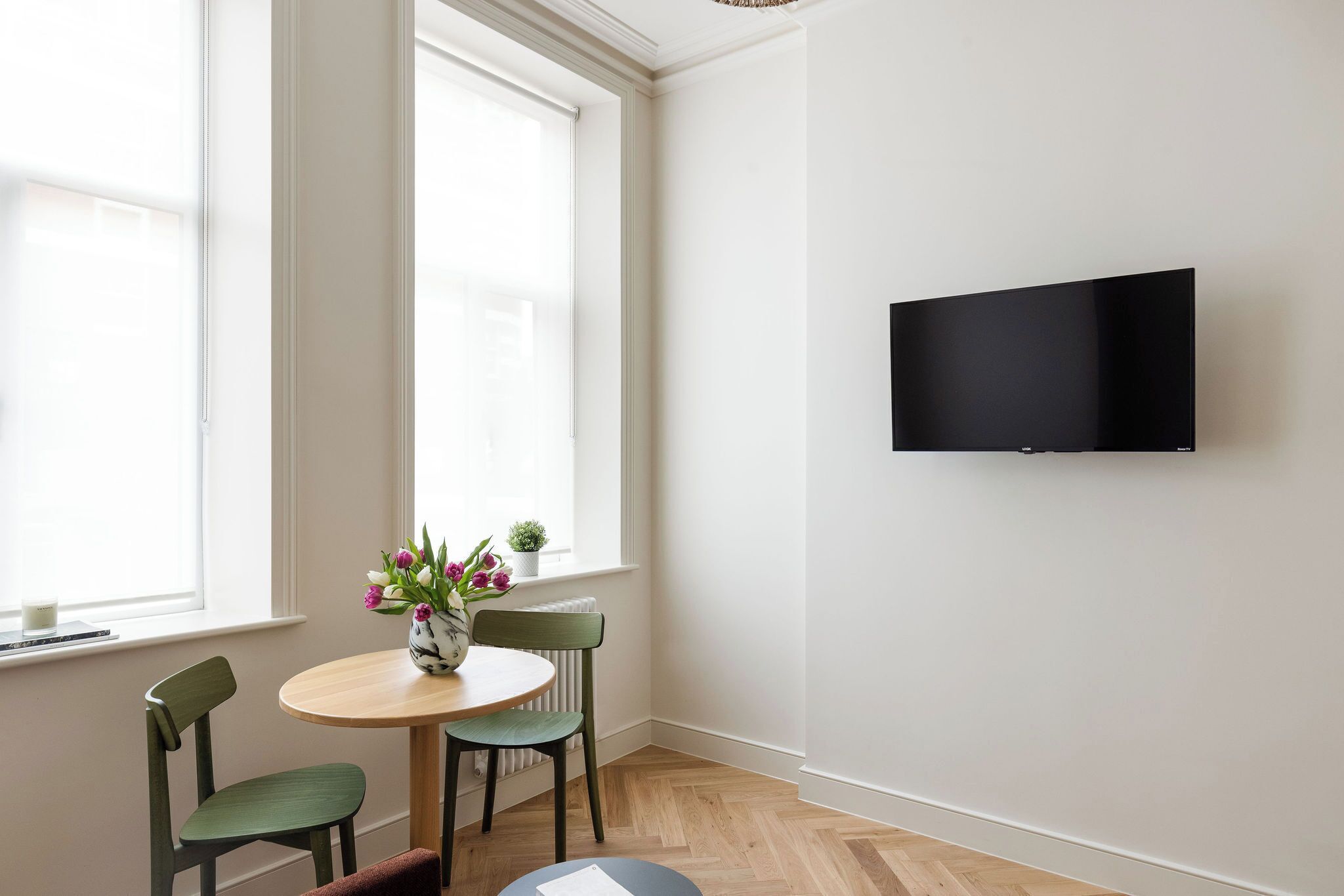 A room with a round table, two chairs, a vase with flowers, a television mounted on the wall, and large windows.