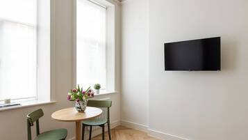 A room with a round table, two chairs, a vase with flowers, a television mounted on the wall, and large windows.
