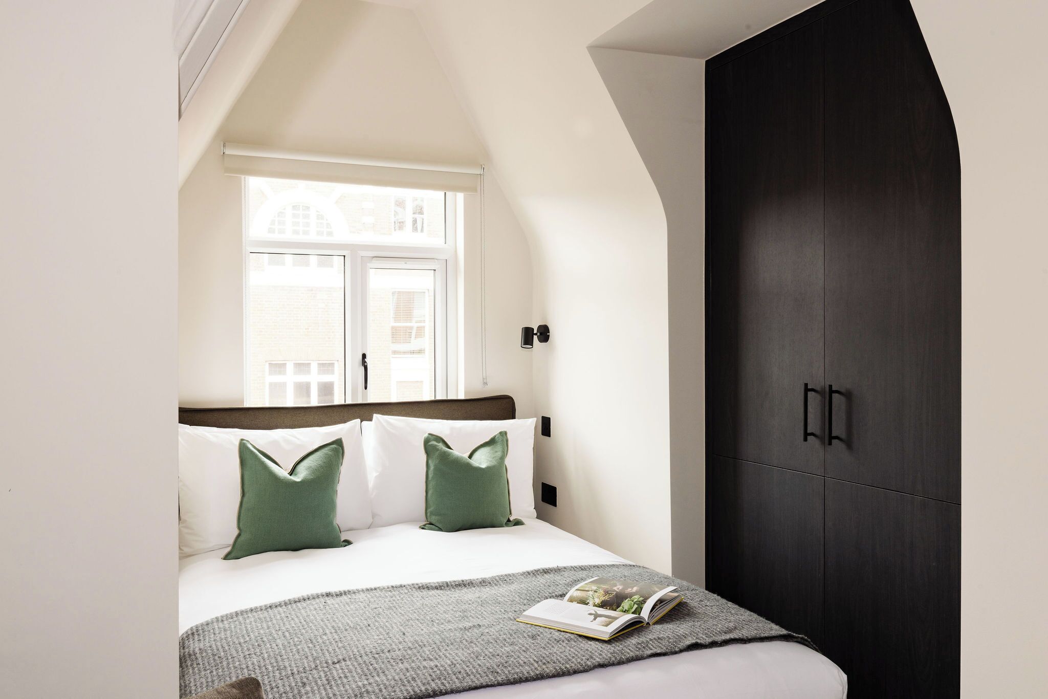 A bedroom with a bed, green pillows, a book, a window, and a dark wardrobe.