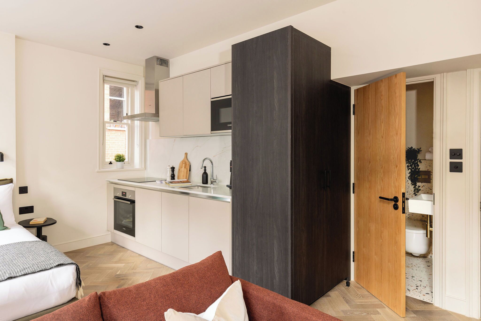 A compact hotel room with a kitchenette, a bed with a red accent pillow, and a wooden door leading to a bathroom.