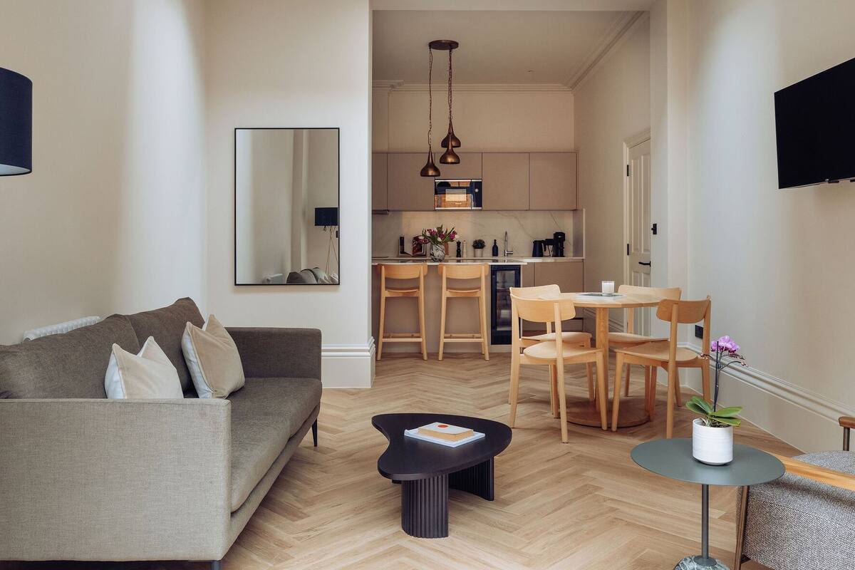 A modern living room with a sofa, a coffee table, and a dining area with chairs and a table. A kitchen is visible in the background with a hanging light fixture.