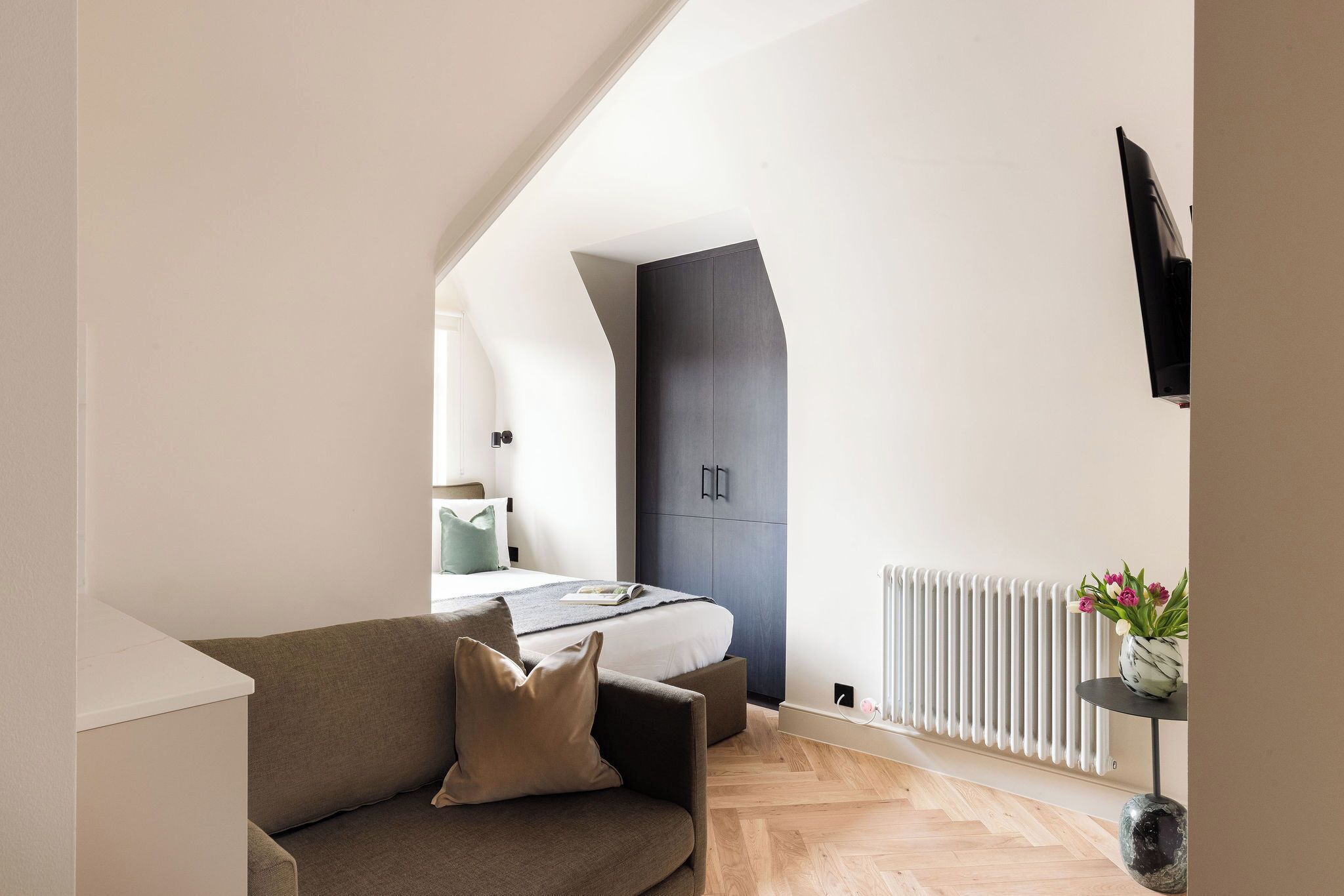 A modern bedroom with a bed, a dark wardrobe, a small table with a vase, and a radiator.