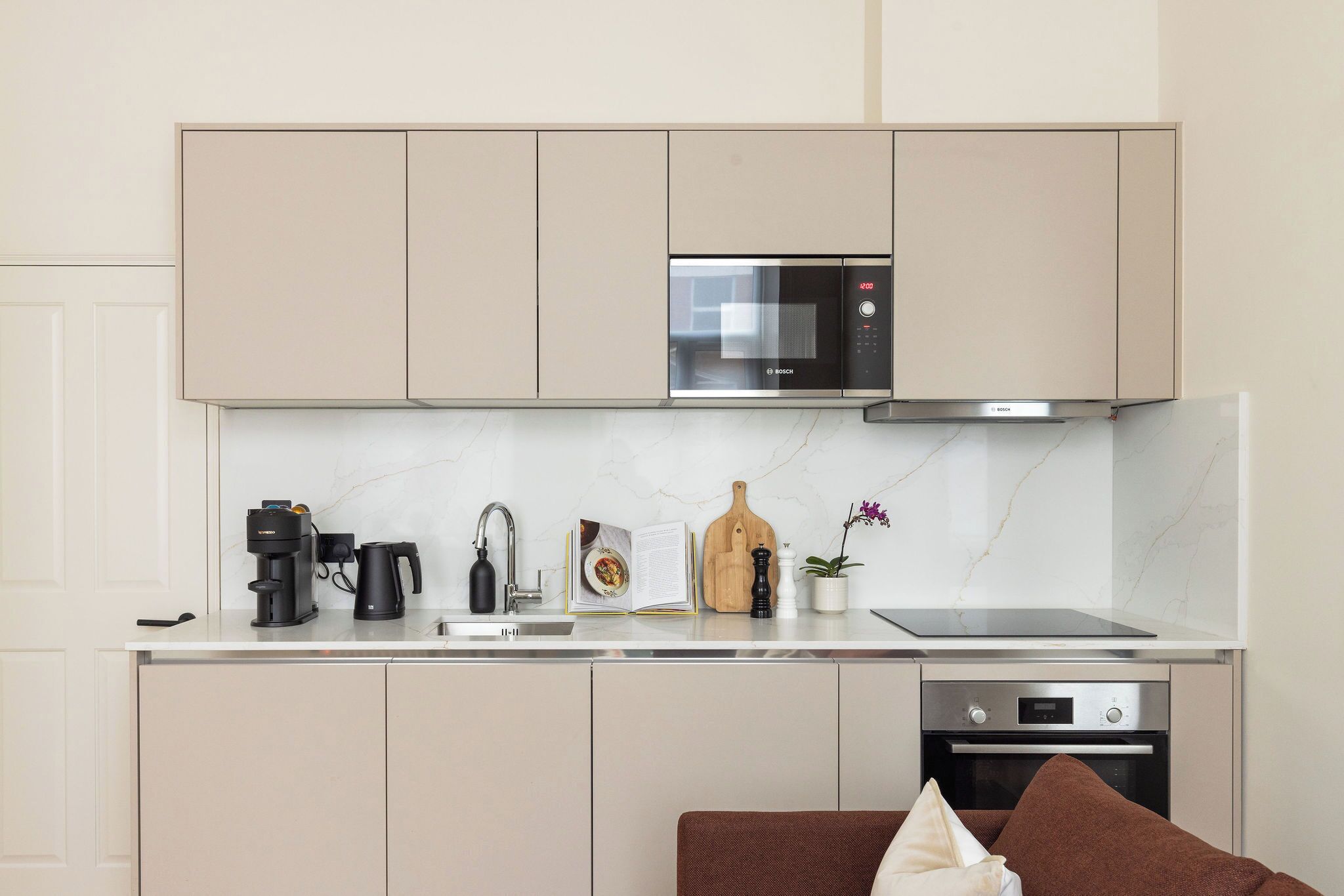 A modern kitchen with beige cabinets, a Bosch microwave, a coffee maker, a kettle, a cutting board, a book, a pepper grinder, and a potted orchid.