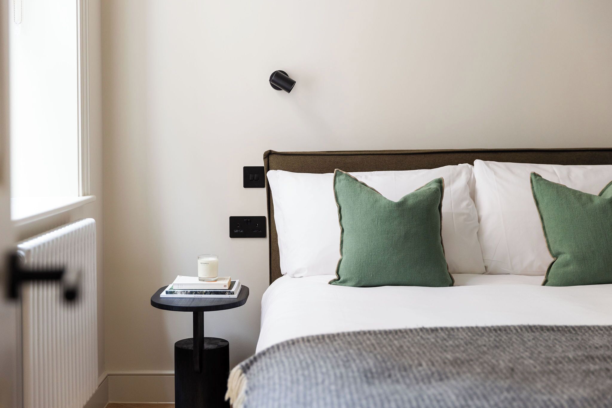Un lit bien fait, orné d’oreillers blancs et verts, une petite table de chevet avec un livre et une bougie, et une lampe fixée au mur.
