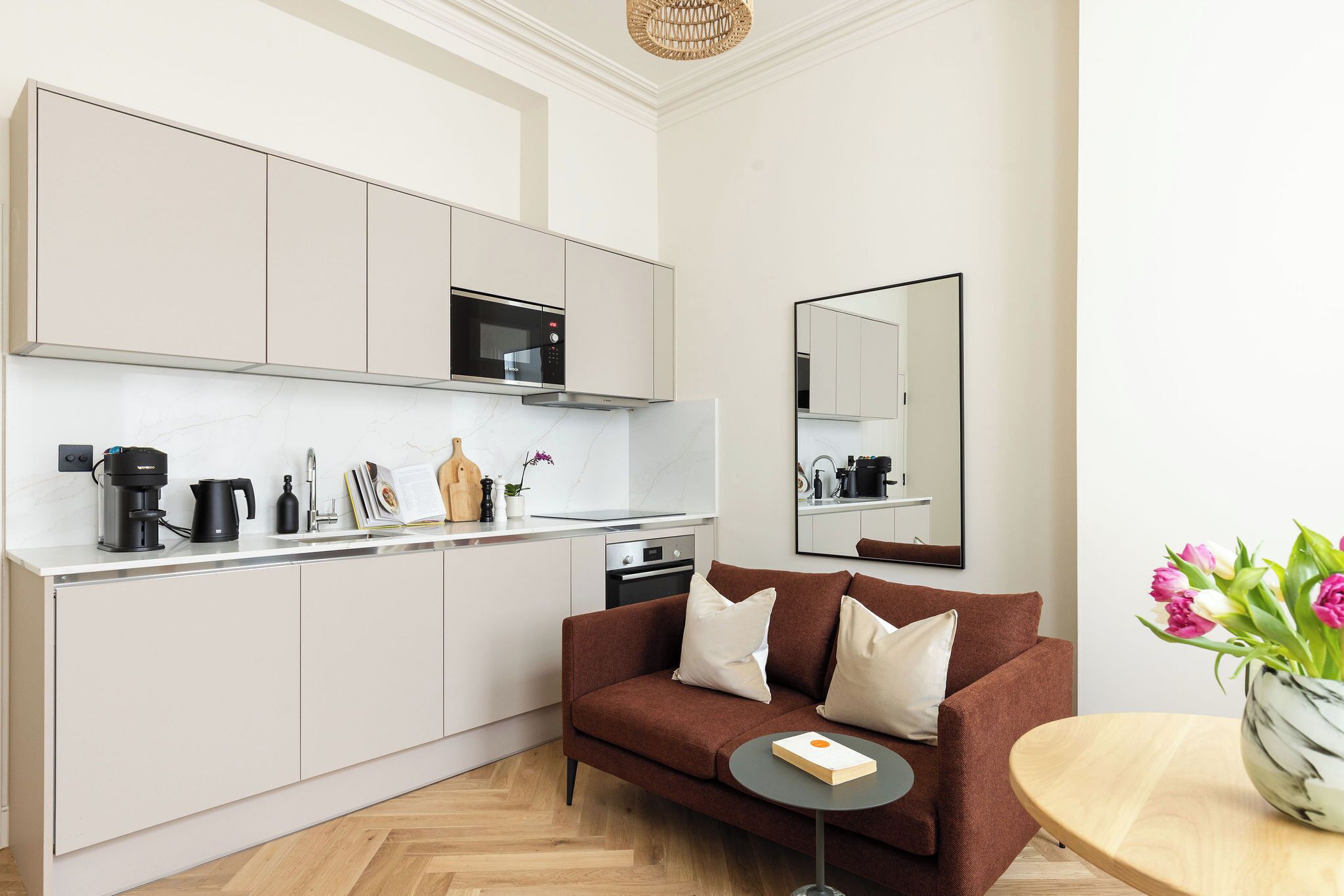 A modern kitchen with white cabinets, a microwave, and a coffee machine. There is a brown sofa with white cushions and a small round table with a book. A vase with flowers is on the table.
