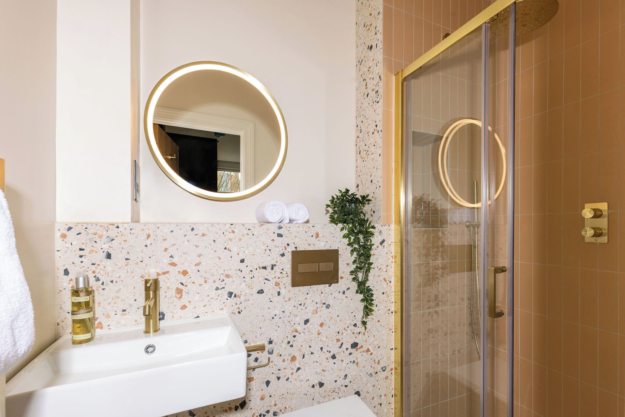 A bathroom with a white sink, gold fixtures, a round mirror with a light, and a shower area with a glass door.