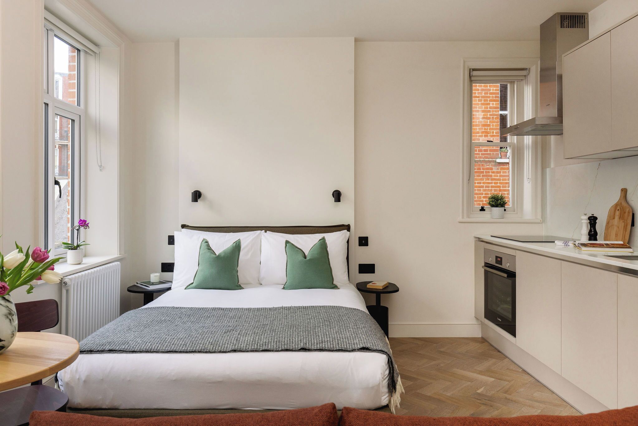 Un dormitorio moderno con una cama grande, una mesita de noche, una pequeña mesita auxiliar, una ventana con vista a un edificio de ladrillo, y una zona de cocina con estufa y armarios.