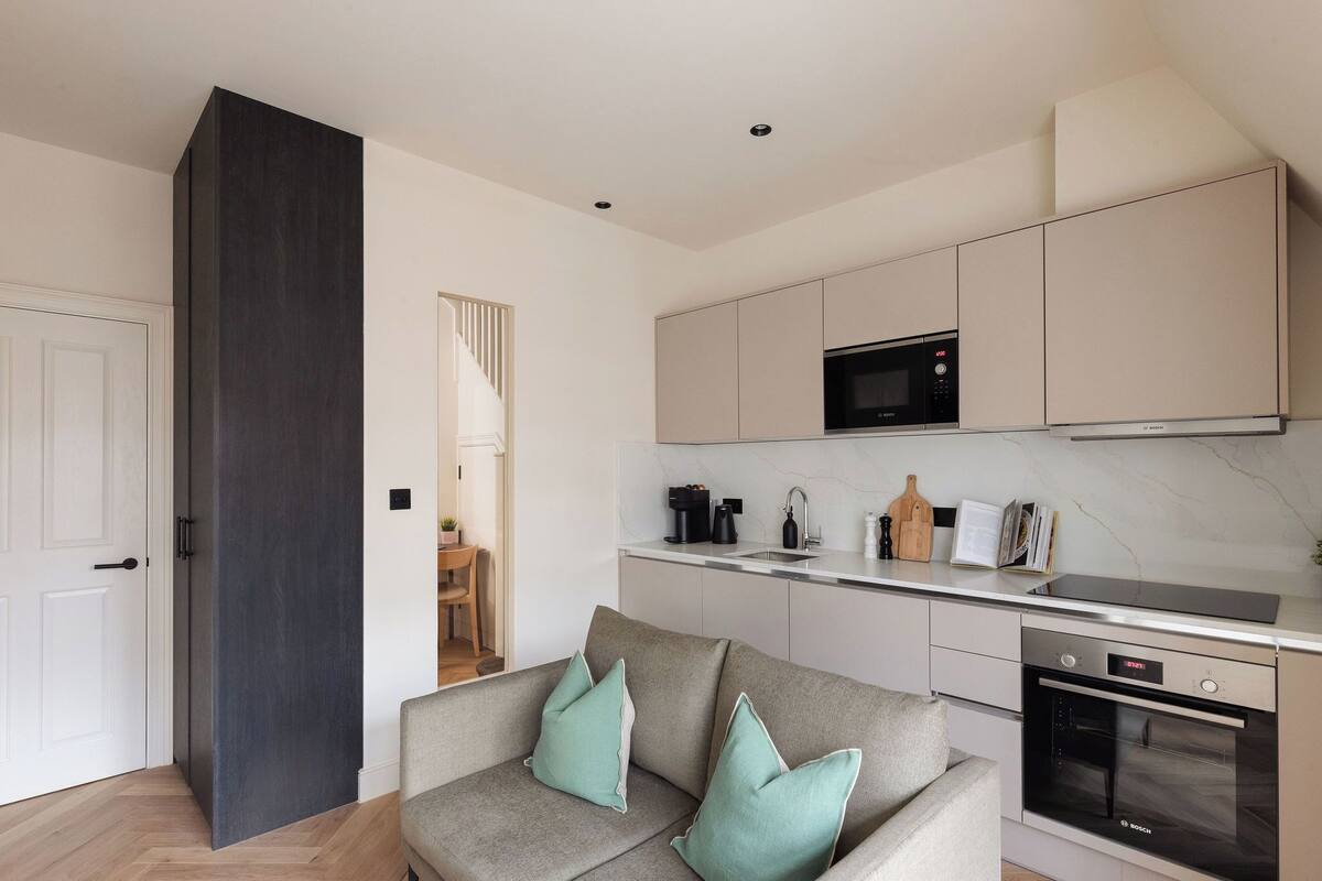 A modern kitchen with a microwave, stove, and oven. There is a grey sofa with teal cushions in the living area.