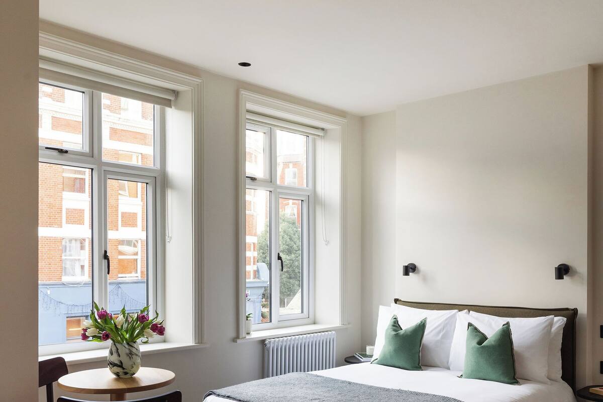 A bedroom with a large bed, a wooden floor, a window with a view of buildings, and a small table with a vase of flowers.
