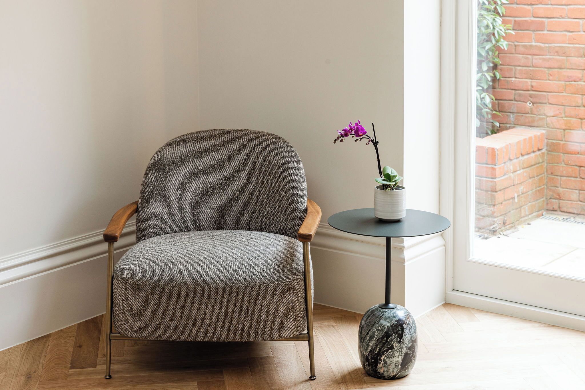 A grey upholstered armchair with wooden armrests and legs, positioned next to a small round table with a marble base, beside a window with a view of a brick wall and greenery.