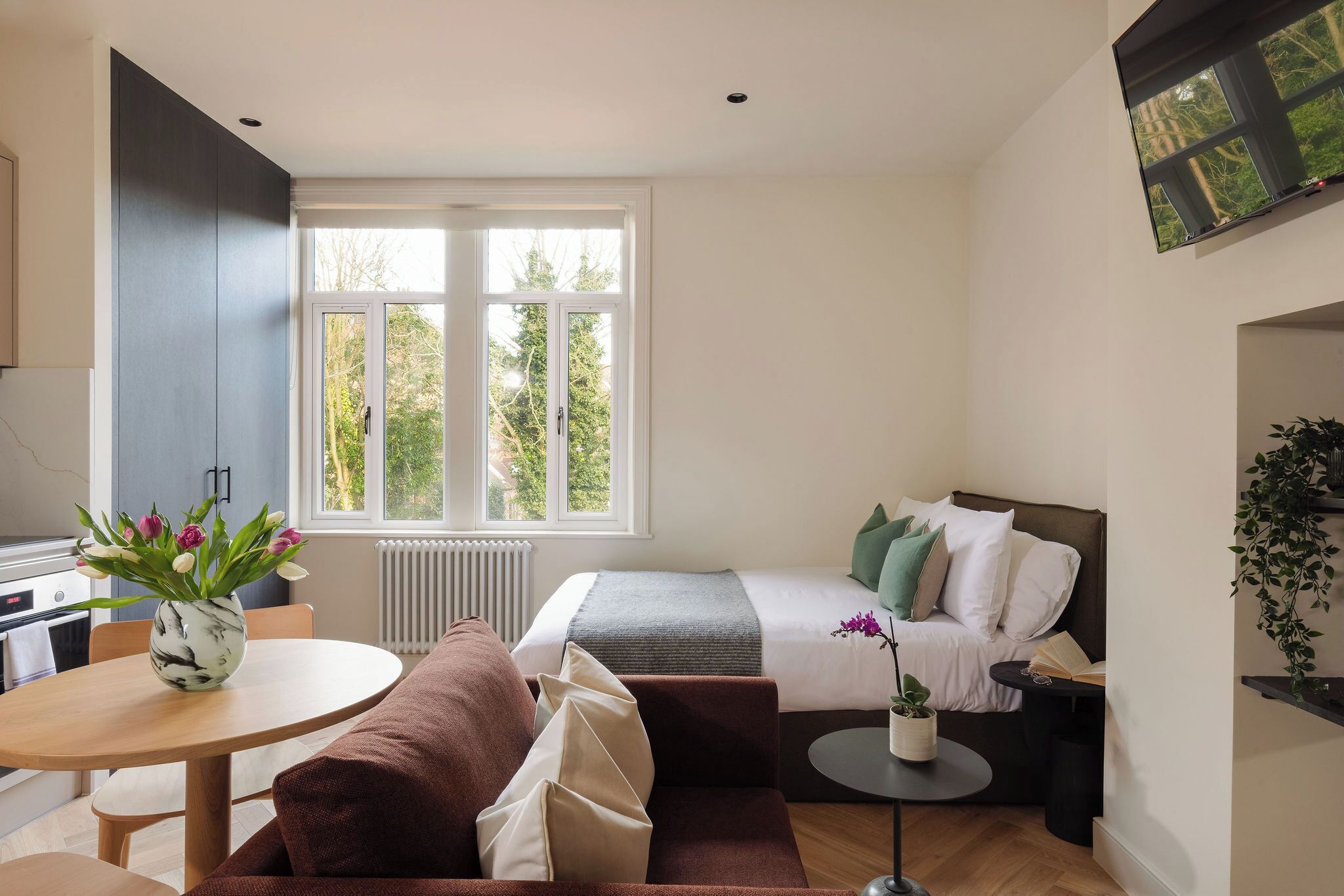 A compact hotel room with a bed, a small round table, a sofa, and a window with a view of greenery.
