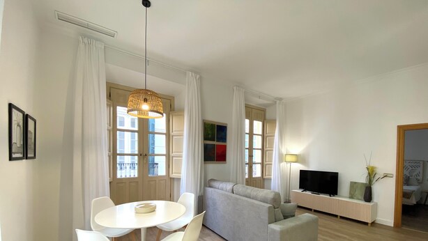 Una sala de estar moderna con un sofá gris, una mesa de comedor blanca con sillas, y un televisor sobre un mueble blanco.