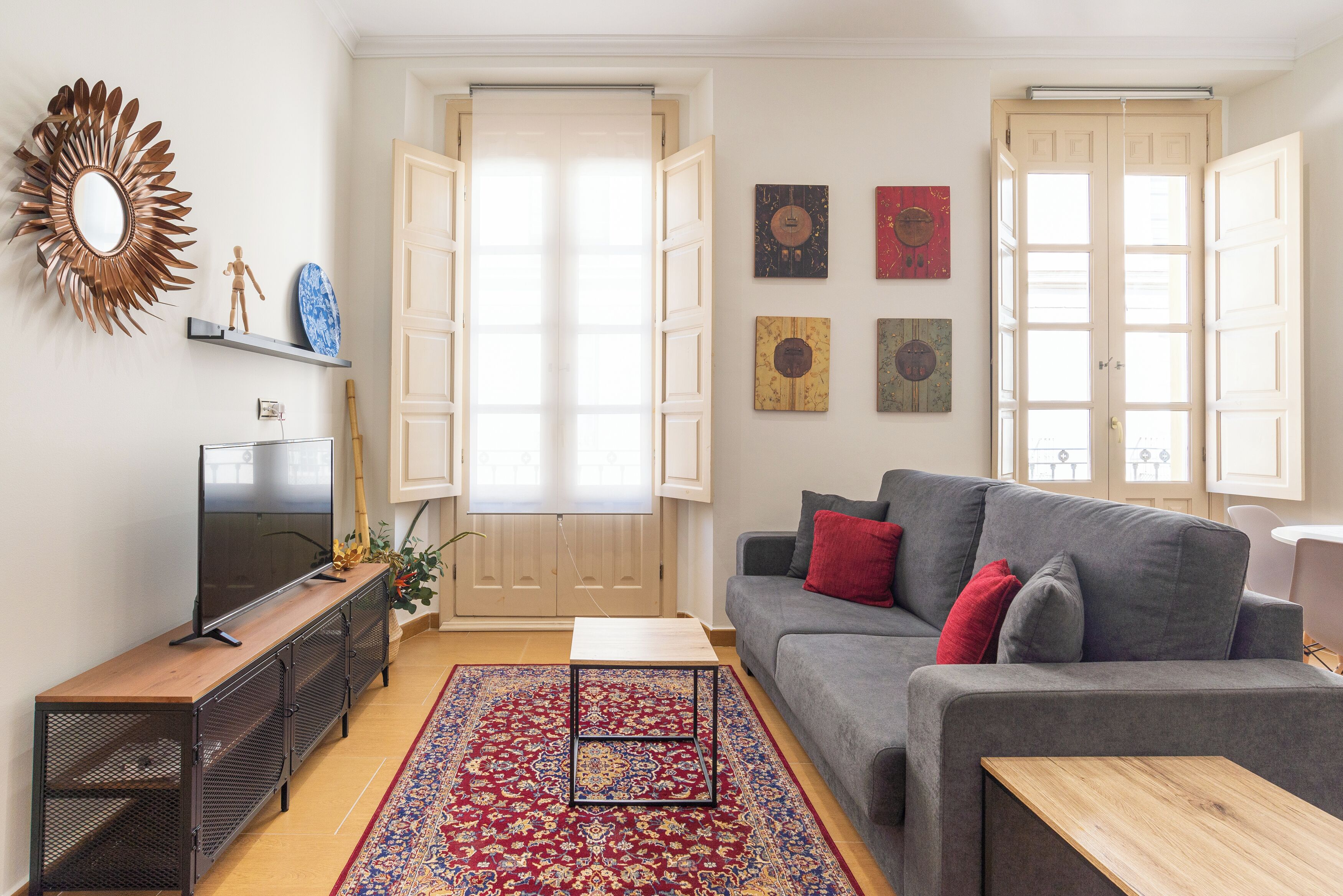 A living room with a grey sofa, red cushions, a wooden coffee table, a TV on a wooden stand, a decorative mirror, and a patterned rug.