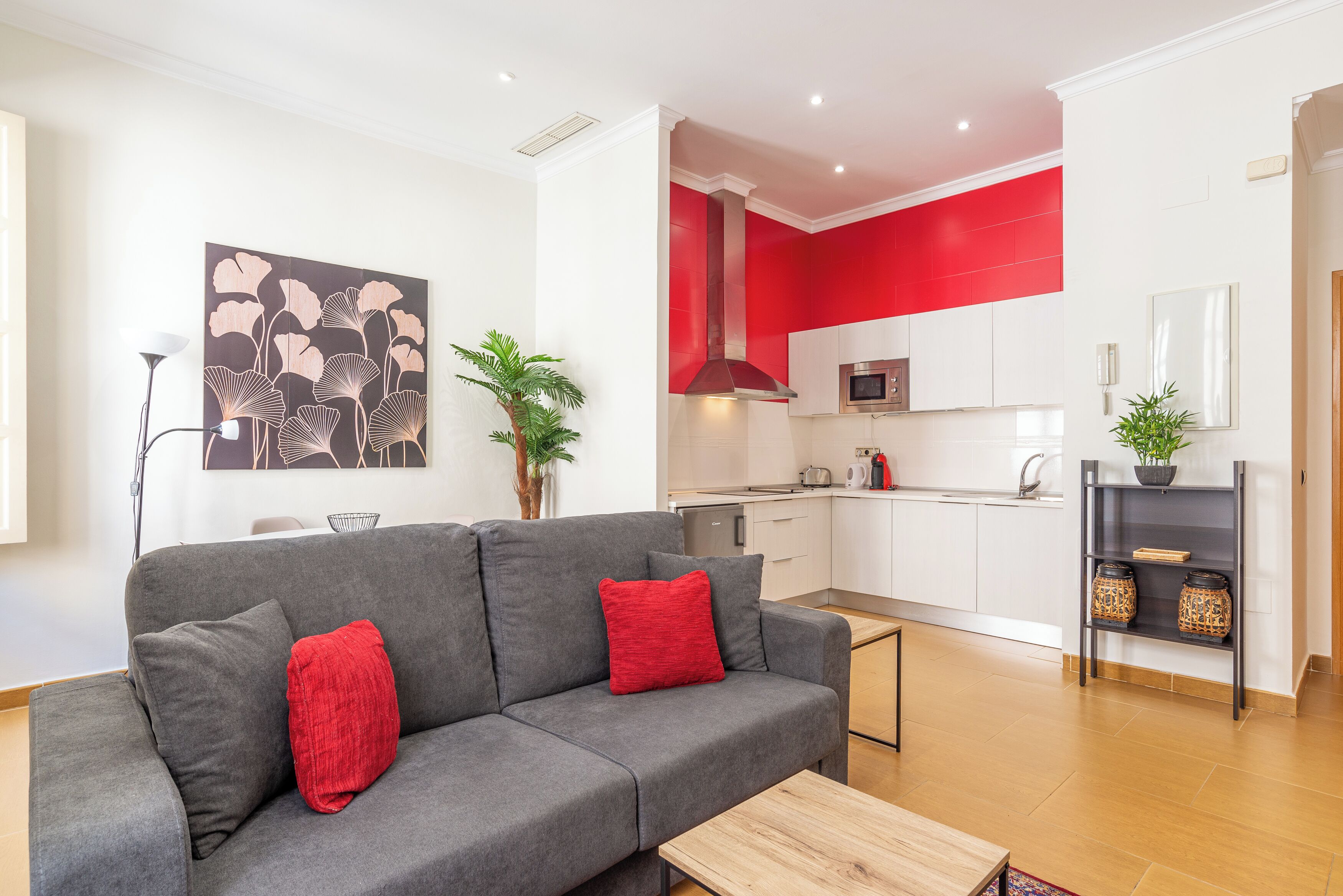 Un salon moderne avec un canapé gris, des coussins rouges et une table basse en bois. On aperçoit, au fond, une cuisine avec des meubles blancs et des touches de rouge.