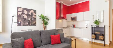 Un salon moderne avec un canapé gris, des coussins rouges et une table basse en bois. On aperçoit, au fond, une cuisine avec des meubles blancs et des touches de rouge.