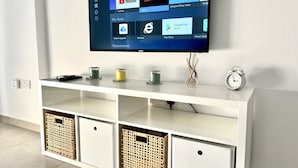 A flat-screen TV mounted on a white wall above a white entertainment center with storage baskets and a clock. - 04 AN - Stay Ease Apartment (Ayia Napa)