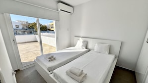 A hotel room with two single beds, a sliding glass door leading to a balcony, and a ceiling air conditioner. - 04 AN - Stay Ease Apartment (Ayia Napa)