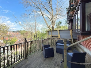 Une terrasse en bois, meublée avec des meubles en osier, dotée d’une rambarde en métal et offrant une vue sur un quartier résidentiel.