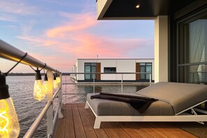 A modern outdoor deck with a lounge area, a view of a modern building across the water, and string lights. - Hausboot am Saaler Bodden, Ribnitz-damgarten (Ribnitz-Damgarten)