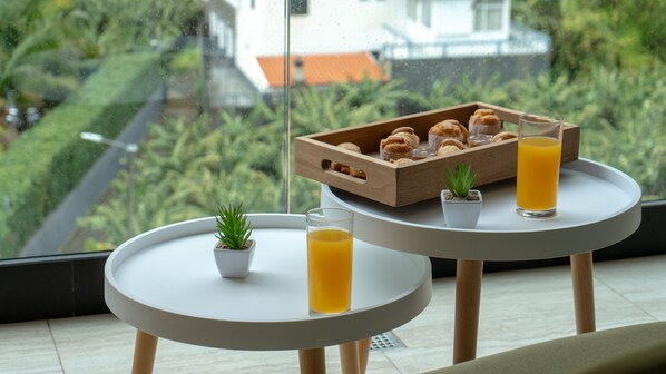 A small table with a tray of pastries and a glass of orange juice, a potted plant, and a view of greenery through a window.
