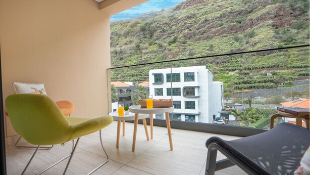 Un balcón con una silla verde, una mesa pequeña con un vaso de jugo de naranja, y vistas a una colina con edificios.