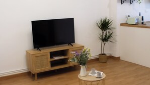 A living room with a flat-screen TV on a wooden cabinet, a small round table with cups and a vase of flowers, and a potted plant.
